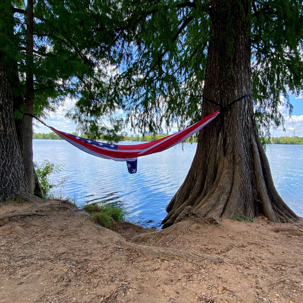 portable hammock with a red, white and blue American flag. Hung up between two large trees on the banks of Lady Bird Lake in Austin, TX